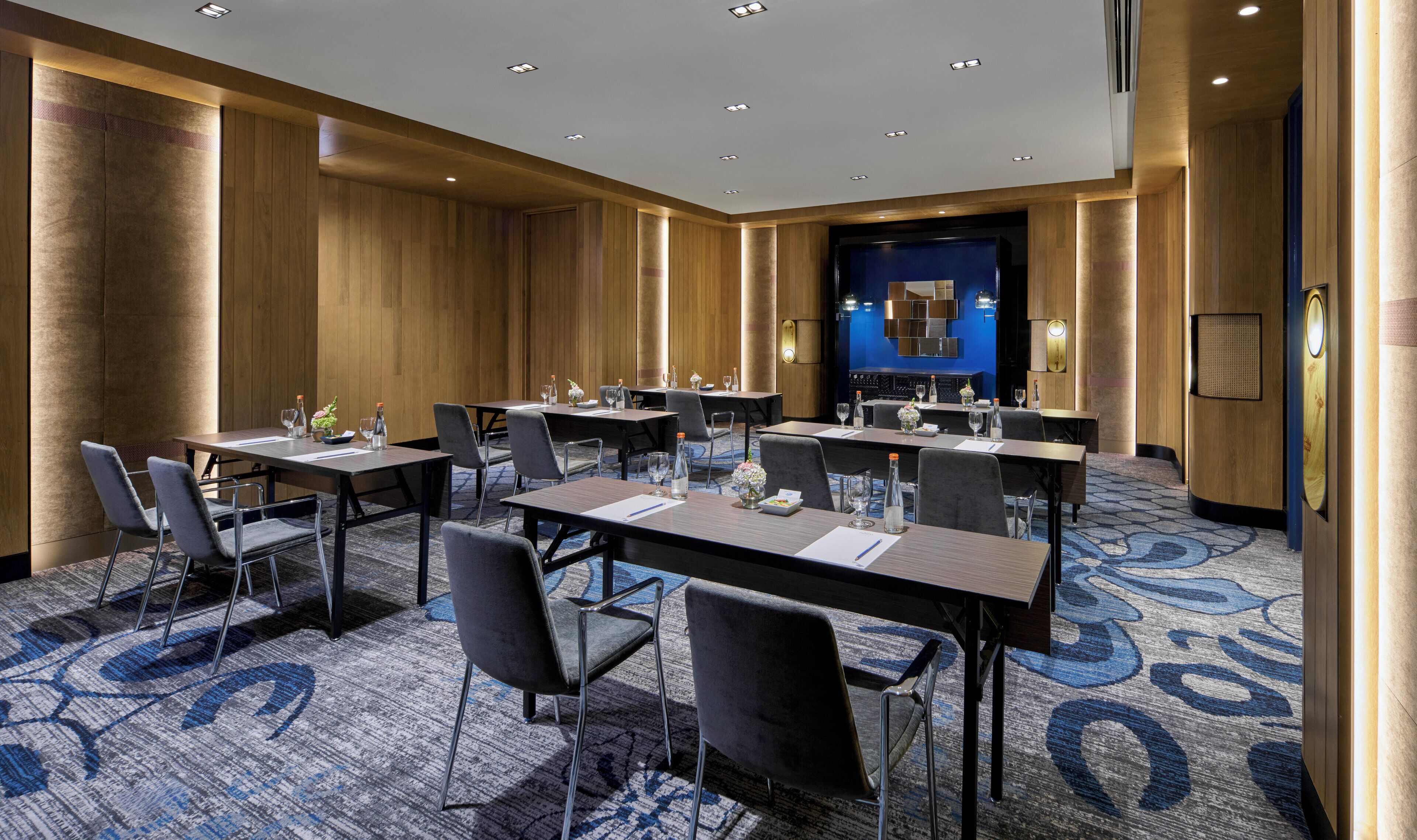 A conference room with multiple tables and chairs, equipped with water bottles and glasses, and a blue carpet with a circular pattern.