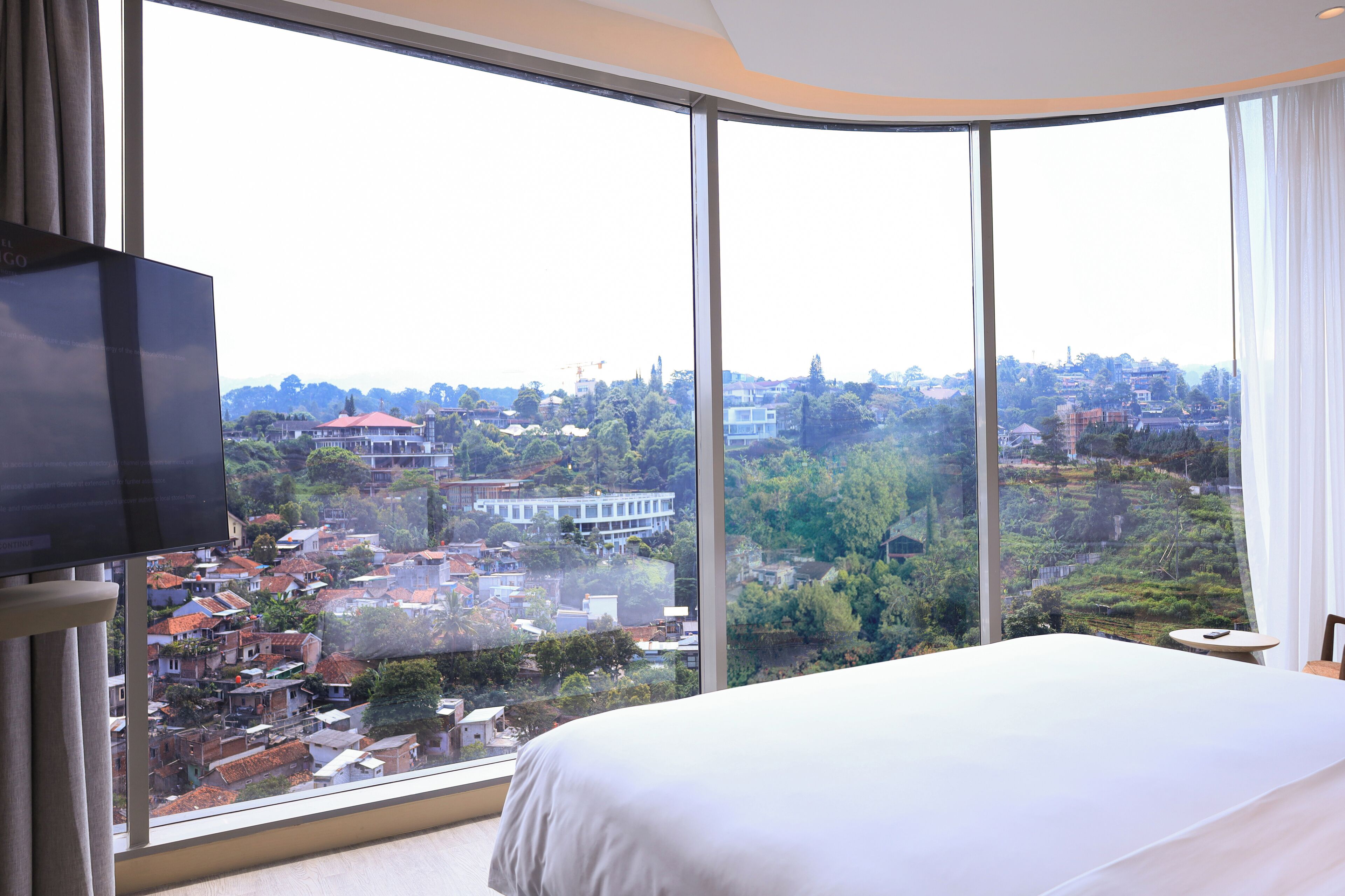 A hotel room with a large window offering a view of a cityscape and greenery.