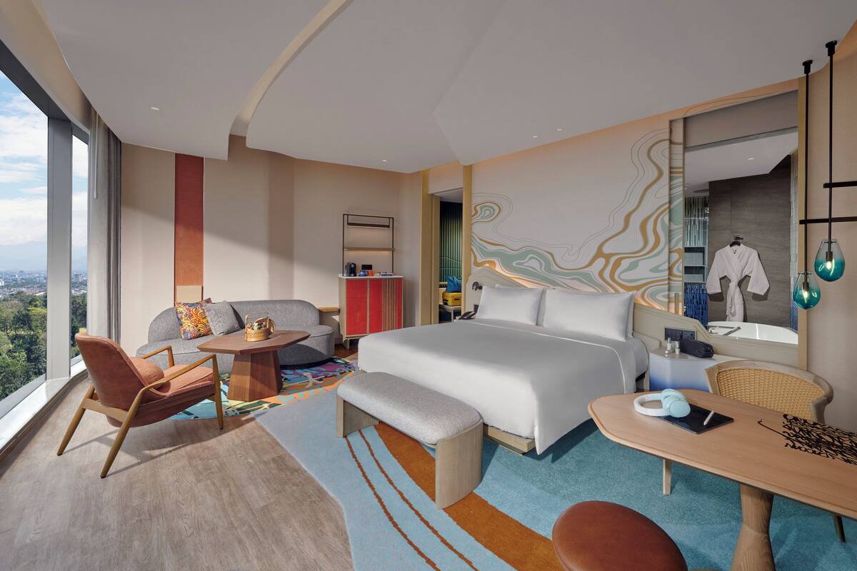 Ett modernt hotellrum med en stor säng, ett skrivbord, en stol och en soffa. Från fönstret har man utsikt över staden.