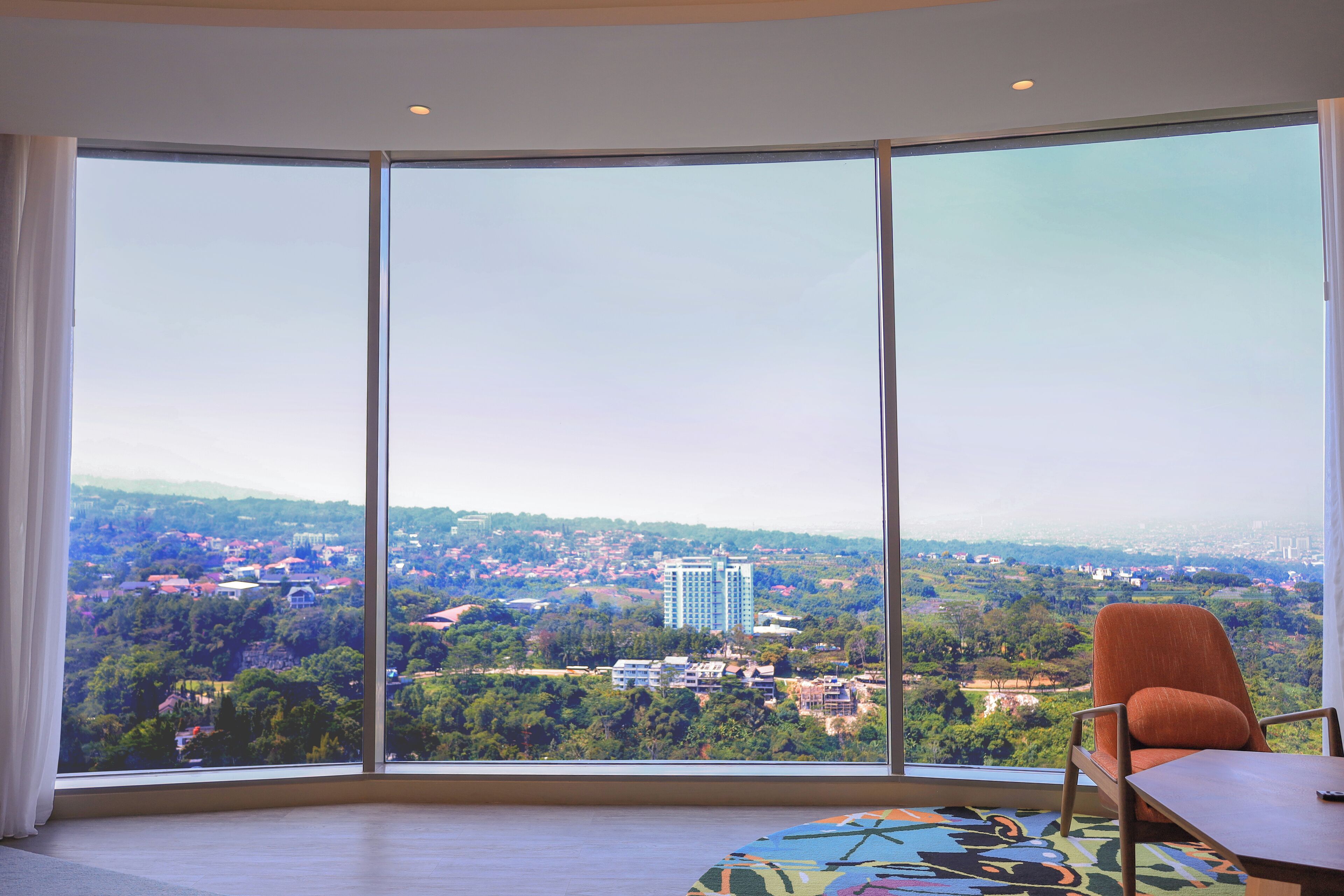 A room with a large window offering a cityscape view, a wooden table, and an orange chair.