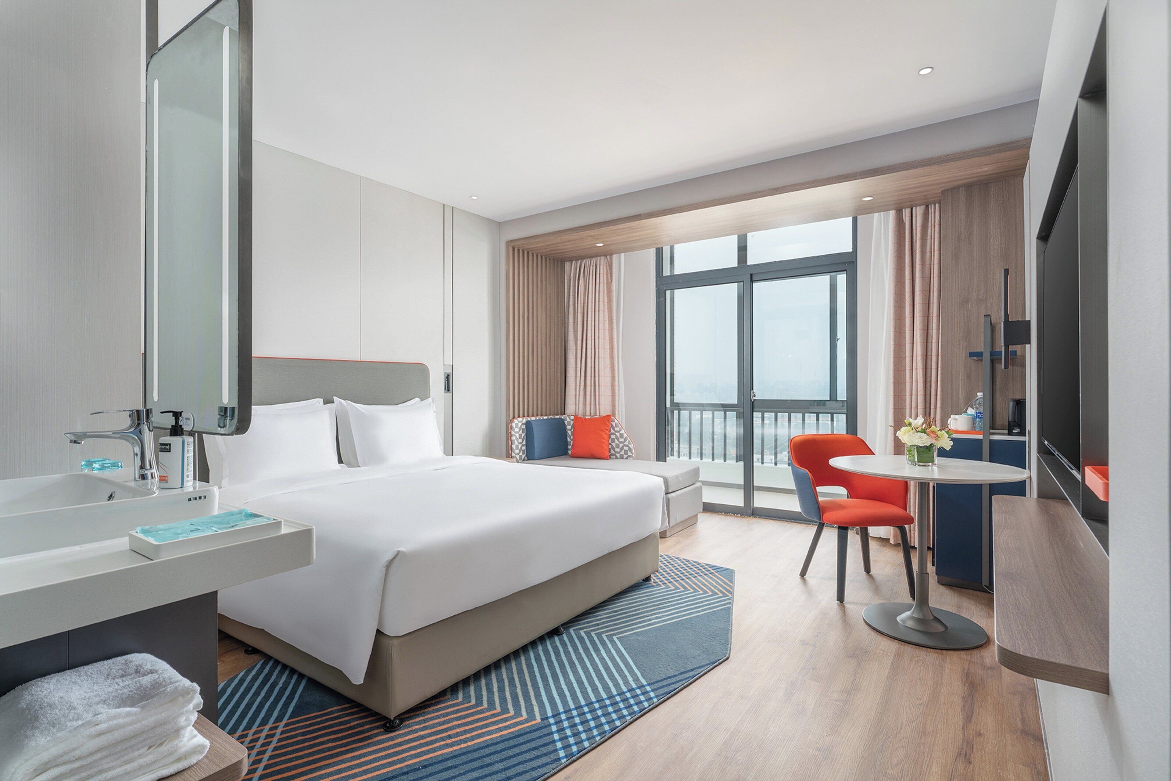 A hotel room with a bed, a desk, a chair, a bathroom, and a view of the city.