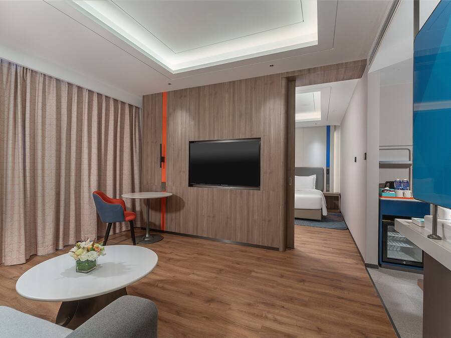 Una moderna camera d'albergo con una TV a schermo piatto, un tavolino rotondo con un vaso di fiori, una poltrona blu e un letto sullo sfondo.