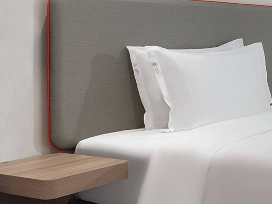 A modern hotel room with a neatly made bed, a wooden nightstand, and a grey headboard with a red trim.