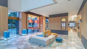 Un hall d’entrée spacieux au design moderne, comprenant un coin salon avec un canapé et un pouf, un bar éclairé par des suspensions rouges, et une grande fenêtre offrant une vue sur l’extérieur.