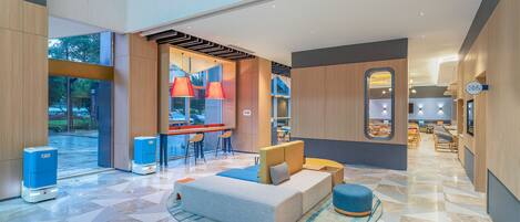 A spacious lobby with a modern design, featuring a seating area with a sofa and ottoman, a bar with red pendant lights, and a large window offering a view of the outdoors.