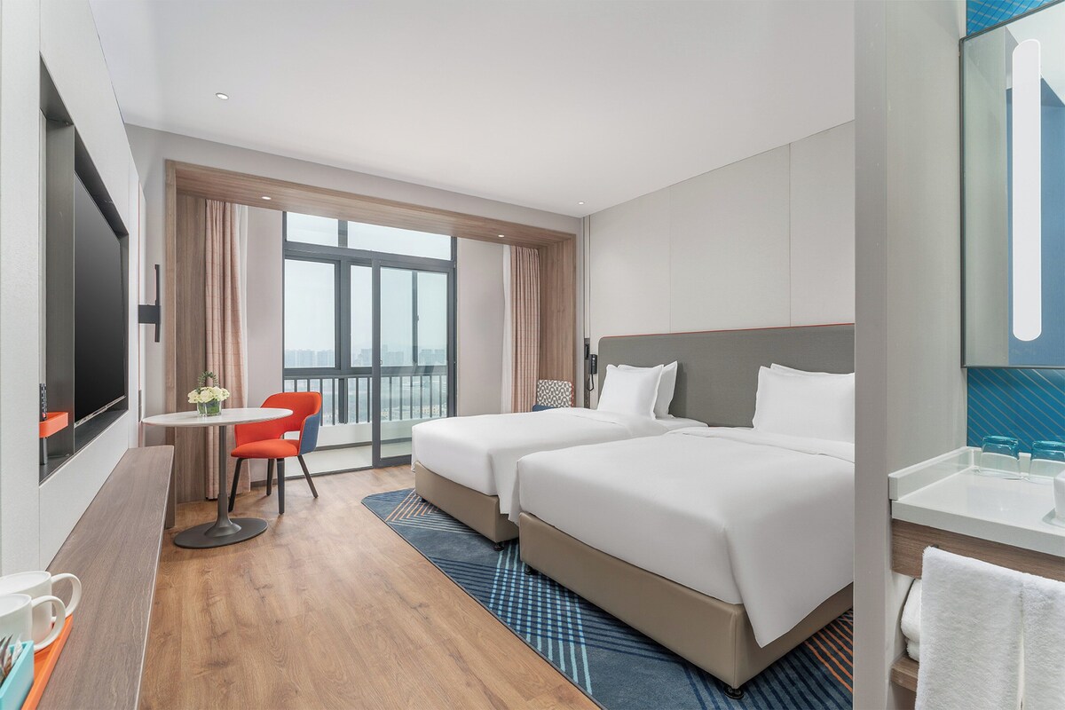 Hotelkamer met twee bedden, een kleine tafel met een stoel, een televisie en een groot raam.
