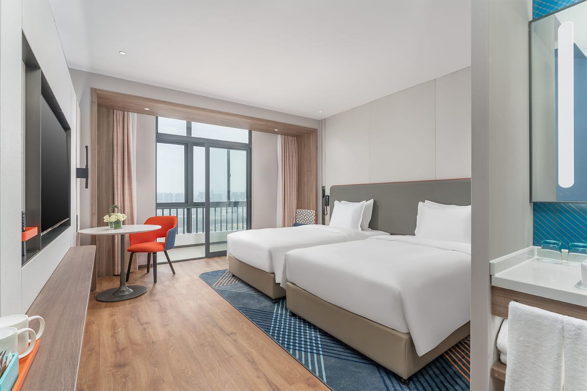 Hotelkamer met twee bedden, een kleine tafel met een stoel, een televisie en een groot raam.