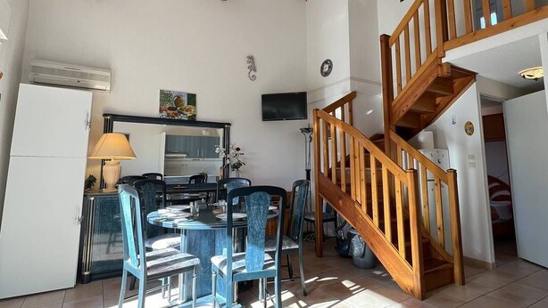 A spacious living area with a dining table, chairs, and a staircase leading to an upper level. - Nice 3 room villa pool Rochelongue Cap d'Agde (LE CAP D AGDE)