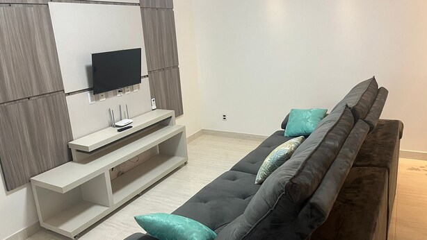 A modern living room with a black sofa, teal cushions, a flat-screen TV, and a white entertainment unit. - Apto DE Luxo Standard 2 Quartos EM Caldas Novas (Olegário Pinto Caldas Novas)