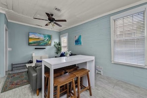 A room with a white desk, two wooden stools, a gray sofa, a ceiling fan, and artwork on the walls.