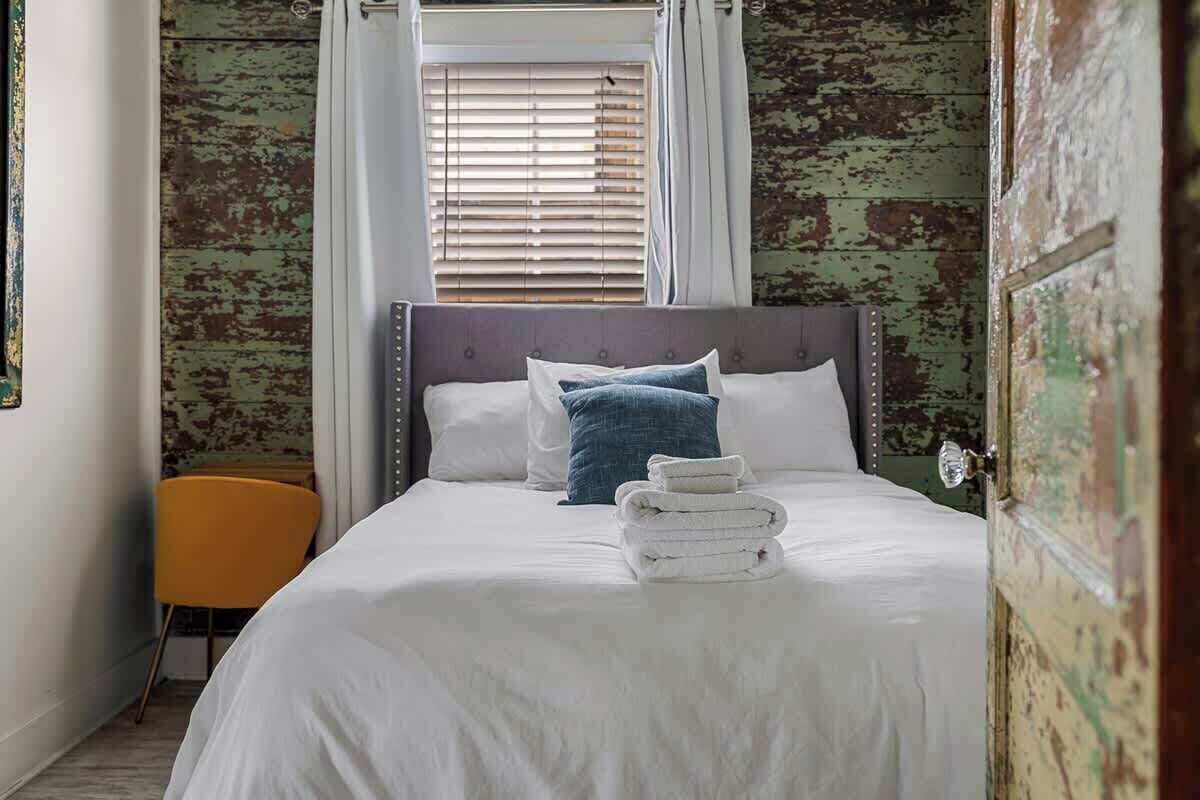 Un lit bien fait, avec des draps blancs et une serviette pliée, une tête de lit rembourrée de couleur grise et un seul oreiller bleu.