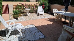 A patio area with white lounge chairs, a table, and a small garden with plants and a palm tree.