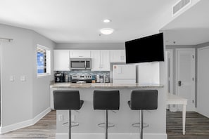 A modern kitchen with white cabinets, a microwave, a coffee maker, and a flat-screen TV mounted above the counter. There are three black bar stools at the counter.