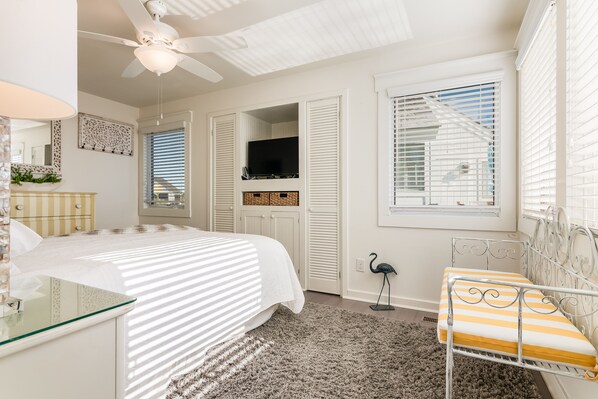 A bedroom with a bed, a bedside table, a mirror, a ceiling fan, a TV, and a window with blinds. - Ocean front villa taking in all views, walking distance to BHI Club; access to both clubs! (Bald Head Island)