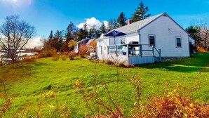 A white single-story house with a porch and a small deck, surrounded by a grassy yard and trees. -  "THE WAY LIFE SHOULD BE"
WATERFRONT OASIS /  HARBOR VIEWS AND A SHARED DOCK.
 (Cushing)