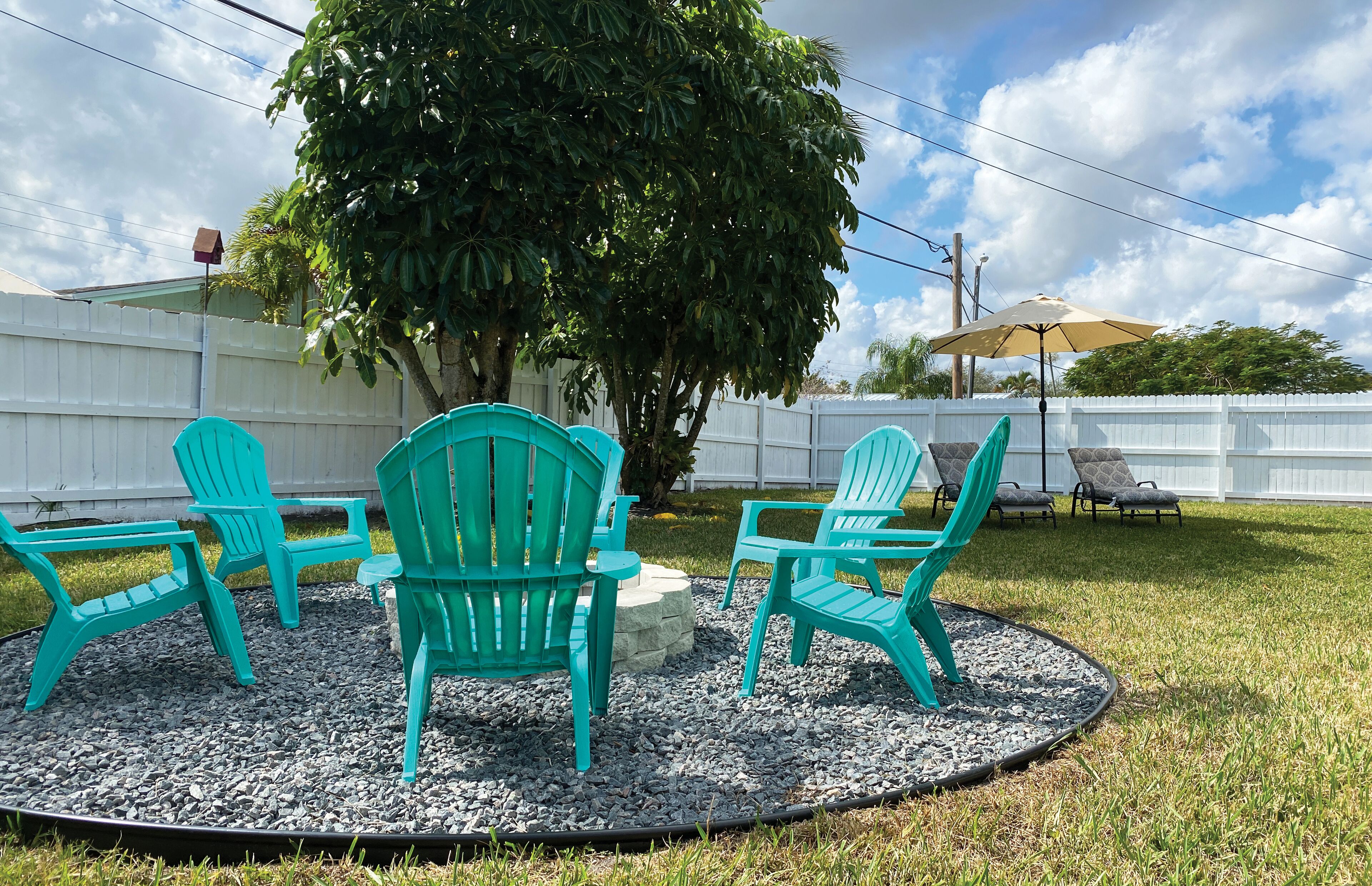 A patio area with four green Adirondack chairs arranged around a central stone feature, a white fence in the background, and a tree providing shade.