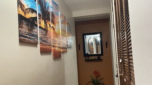 A hallway with three wall-mounted paintings depicting tropical scenes, a potted plant, and a door with a decorative panel.