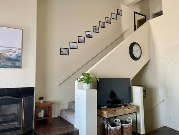 A staircase with a clock and framed photos on the wall, a television on a wooden stand, and a fireplace in the foreground.