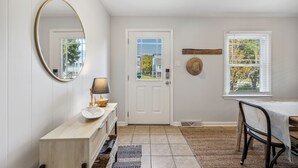 Un couloir avec une porte blanche, un miroir rond, une console avec une lampe, une table à manger avec des chaises et une fenêtre avec des stores.