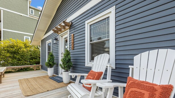 Une terrasse en bois avec des chaises Adirondack blanches, une petite plante en pot et une plaque numérotée sur la maison.