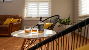 Een ronde tafel met een fles en twee glazen, een bruine leren bank met gele kussens, en een zwarte rieten stoel.