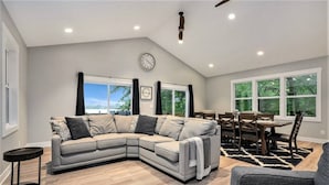 A modern living room with a sectional sofa, a dining table with chairs, a clock on the wall, and large windows with a view. - Tranquil Jewel: Arcade, King Suits, Hot Tub, Decks (Sand Lake)