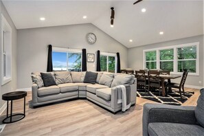 A modern living room with a sectional sofa, a dining table with chairs, a clock on the wall, and large windows with a view. - Tranquil Jewel: Arcade, King Suits, Hot Tub, Decks (Sand Lake)