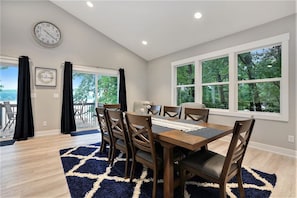 A dining area with a wooden table, chairs, a clock, and large windows with a view of trees. - Tranquil Jewel: Arcade, King Suits, Hot Tub, Decks (Sand Lake)