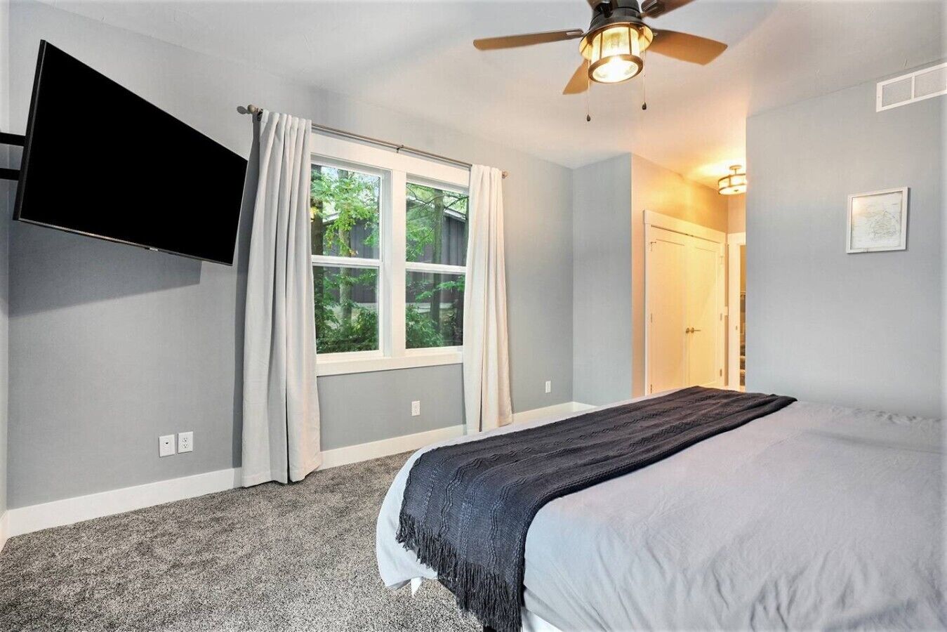 A bedroom with a bed, a ceiling fan, a TV mounted on the wall, a window with curtains, and a door leading to another room.
