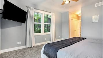 A bedroom with a bed, a ceiling fan, a TV mounted on the wall, a window with curtains, and a door leading to another room.