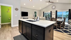 A modern kitchen with a central island, a dining area with a table and chairs, a living room with a sofa, and a balcony with a view. - Tranquil Jewel: Arcade, King Suits, Hot Tub, Decks (Sand Lake)