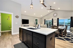 A modern kitchen with a central island, a dining area with a table and chairs, a living room with a sofa, and a balcony with a view. - Tranquil Jewel: Arcade, King Suits, Hot Tub, Decks (Sand Lake)