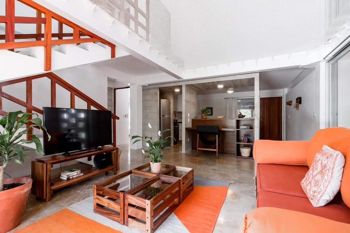 Amplia sala de estar con televisor, mesa de centro de madera, sofá naranja y plantas en macetas.