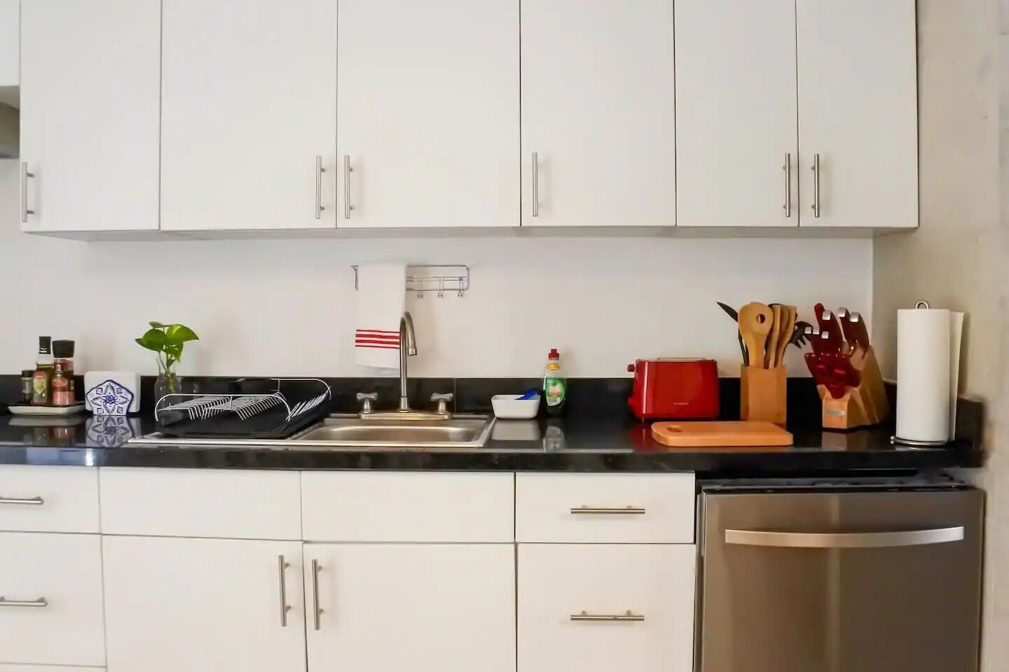 A kitchen with white cabinets, a stainless steel sink, and a red toaster. There are various kitchen utensils and a plant on the counter.