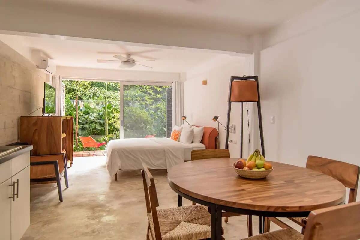 Una habitación moderna con cama, una mesa de comedor con fruta, un ventilador de techo y vistas a zonas verdes.