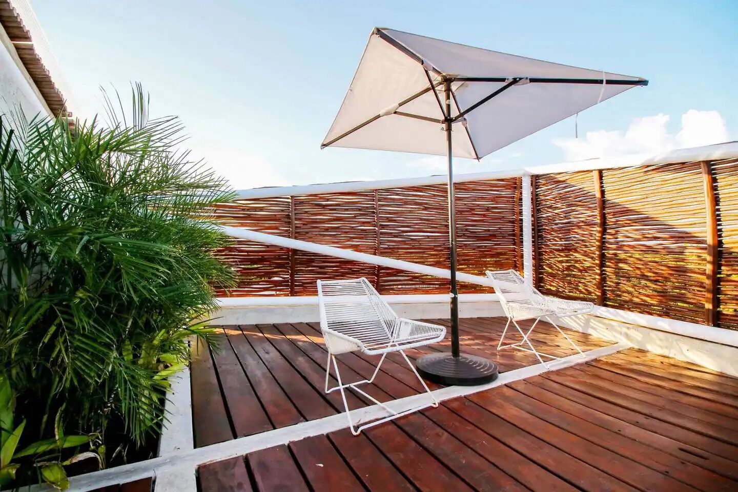 A wooden deck with a white metal chair and a large umbrella, surrounded by lush greenery and a bamboo fence.