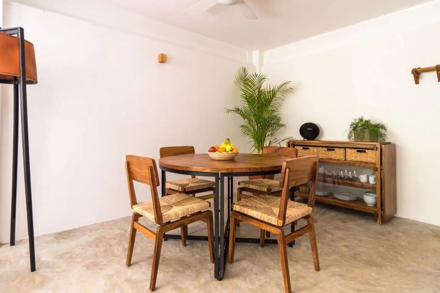 A dining area with a round wooden table, six woven chairs, a fruit basket, a potted plant, and a wooden cabinet with glass doors.
