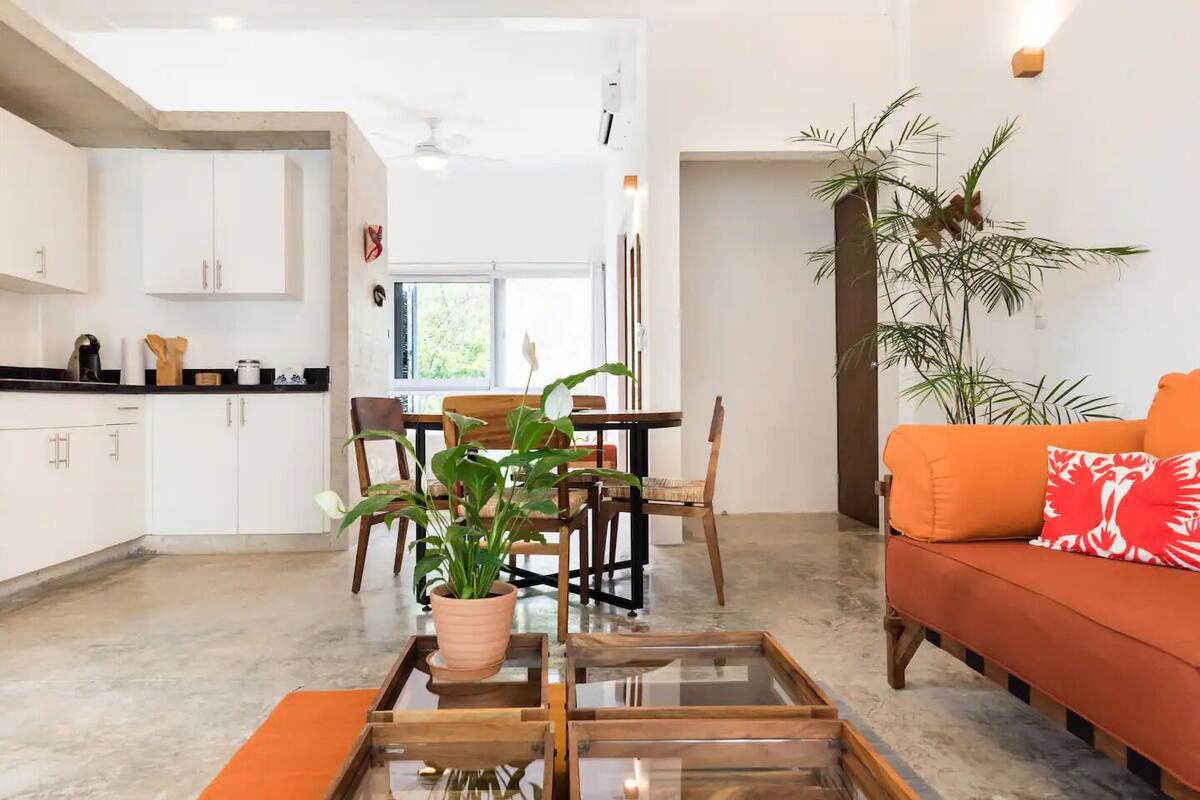 Un espacio habitable moderno con zona de comedor, sofá con cojines naranja y una planta en maceta.