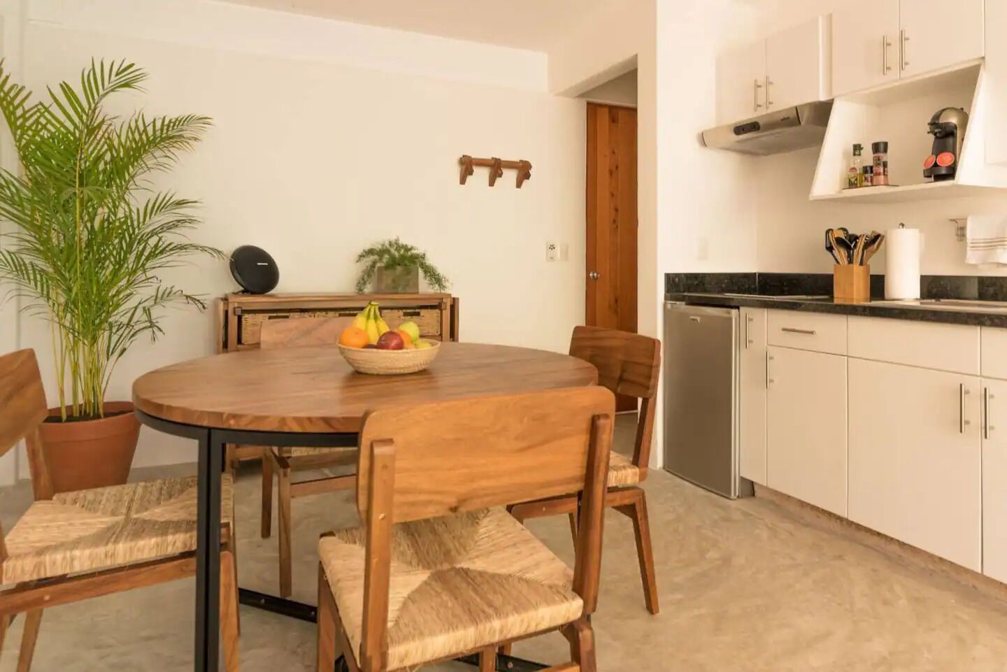 A dining area with a round wooden table, four chairs, a potted plant, a fruit bowl, and a kitchen area with cabinets and appliances.