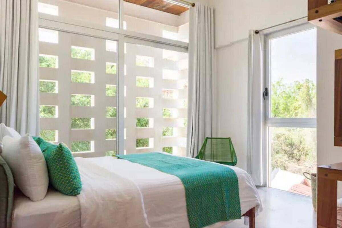 Un dormitorio con una cama, una almohada verde, una almohada blanca, una manta verde, una mesita de noche de madera y una ventana con vistas a la vegetación.