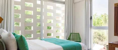 Un dormitorio con una cama, una almohada verde, una almohada blanca, una manta verde, una mesita de noche de madera y una ventana con vistas a la vegetación.