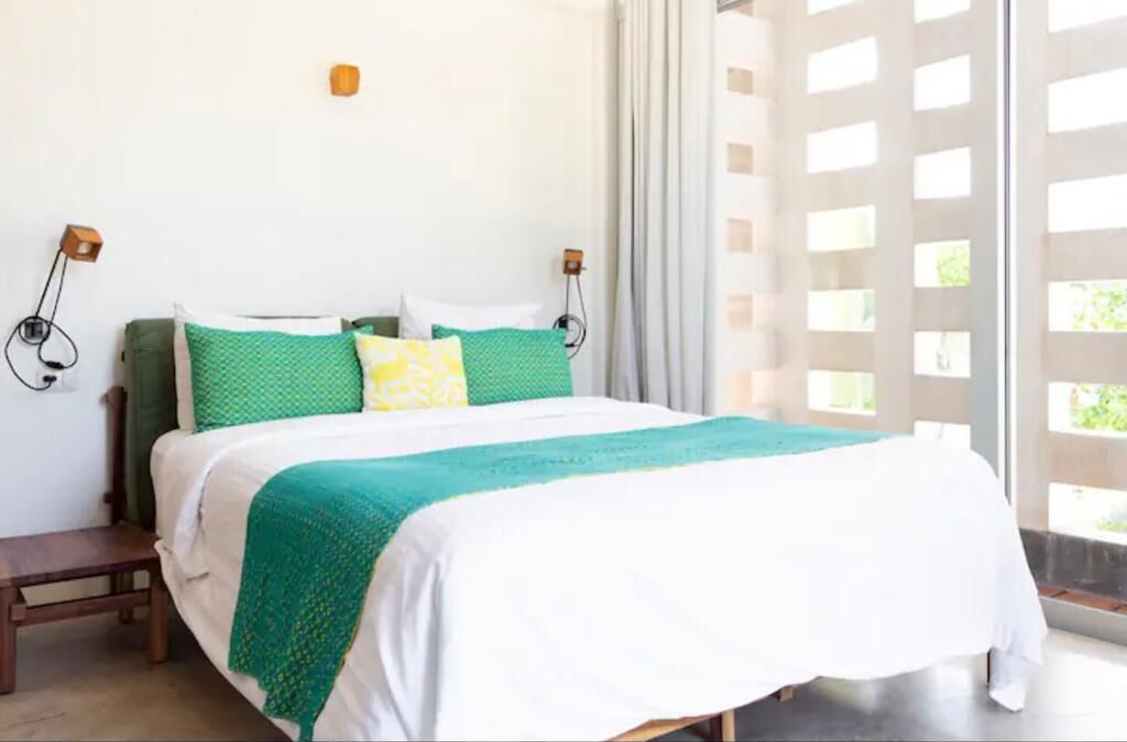 A bed with a green and white bedspread, two pillows, a wooden bench, and a wall-mounted light fixture.