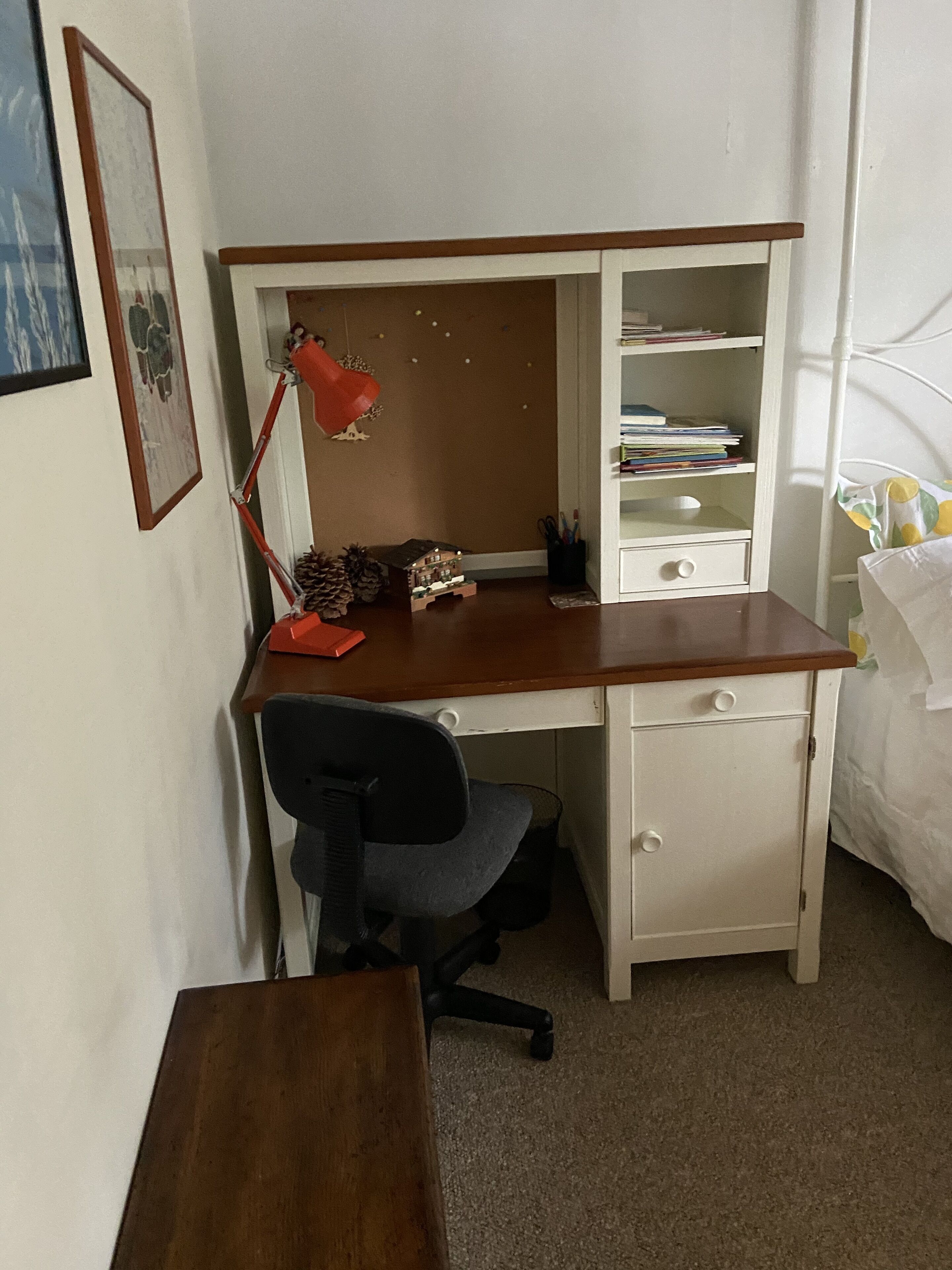A compact bedroom with a wooden desk, a chair, a bed with a white and yellow patterned bedspread, and a bookshelf with books and decorative items.