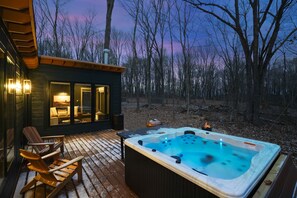 A wooden deck with a hot tub and Adirondack chairs, overlooking a forested area.