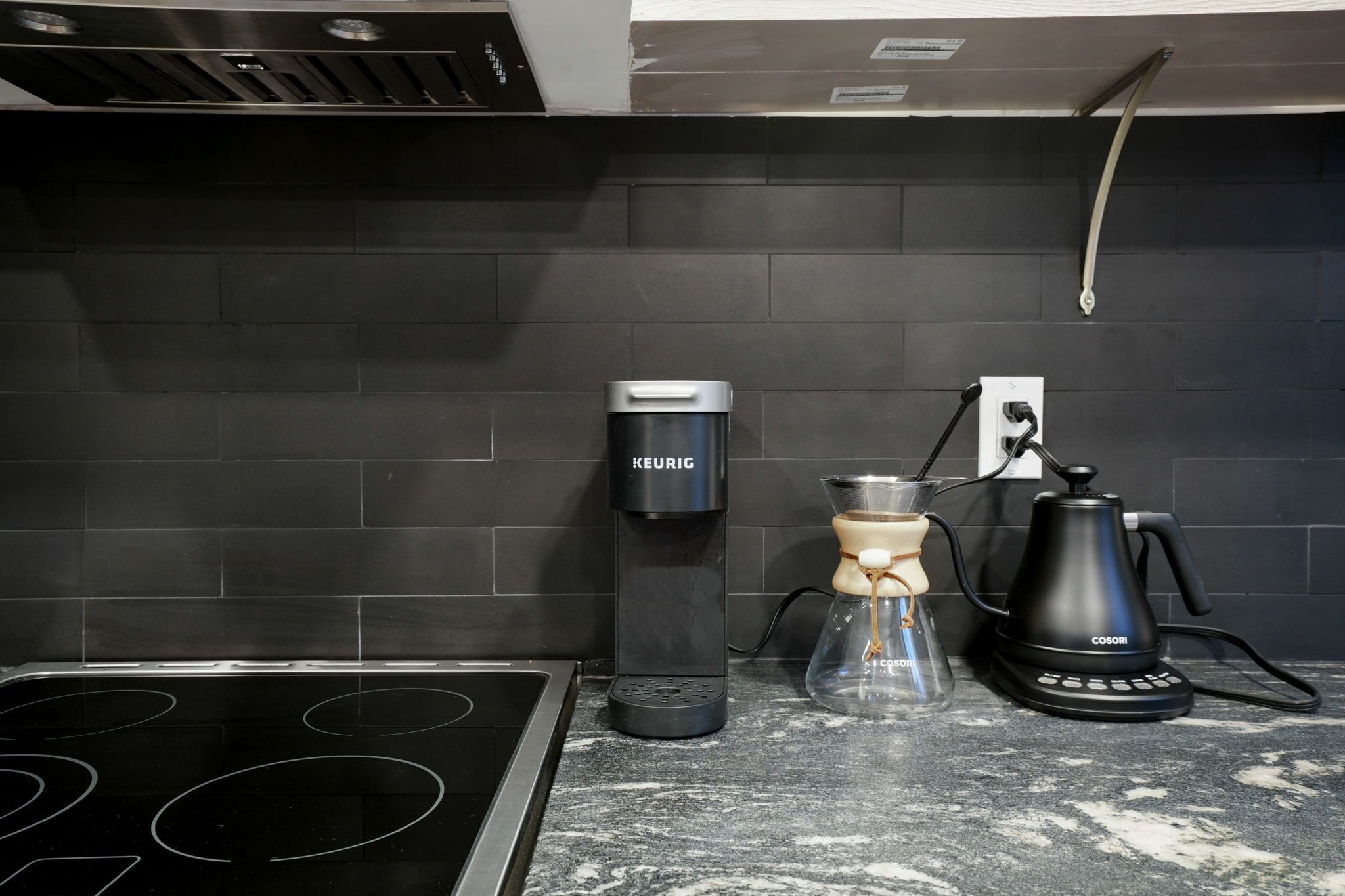 A kitchen with a black tiled backsplash, a Keurig coffee maker, a glass carafe, and a Cosori coffee pot.