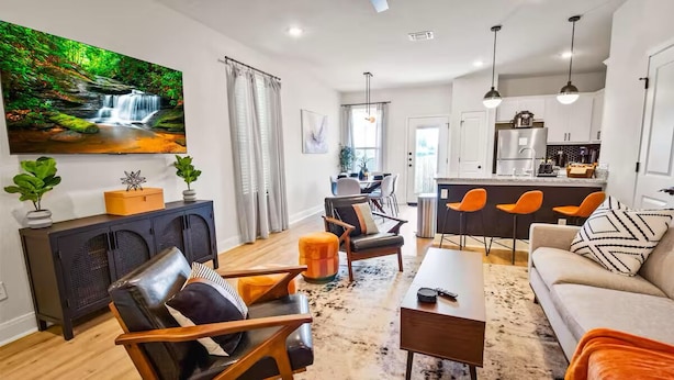 Un salon moderne comprenant un canapé, des fauteuils, une table basse et un téléviseur. Il y a également un coin cuisine avec une table à manger et des chaises.