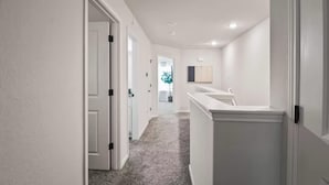 Un couloir aux murs blancs, doté d’un sol recouvert de moquette et d’une porte donnant sur une salle de bain équipée d’un lavabo et d’un miroir.