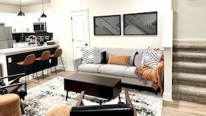 Un salon moderne avec un canapé, une table basse et des fauteuils. On aperçoit une cuisine au fond, avec un four à micro-ondes et des placards.