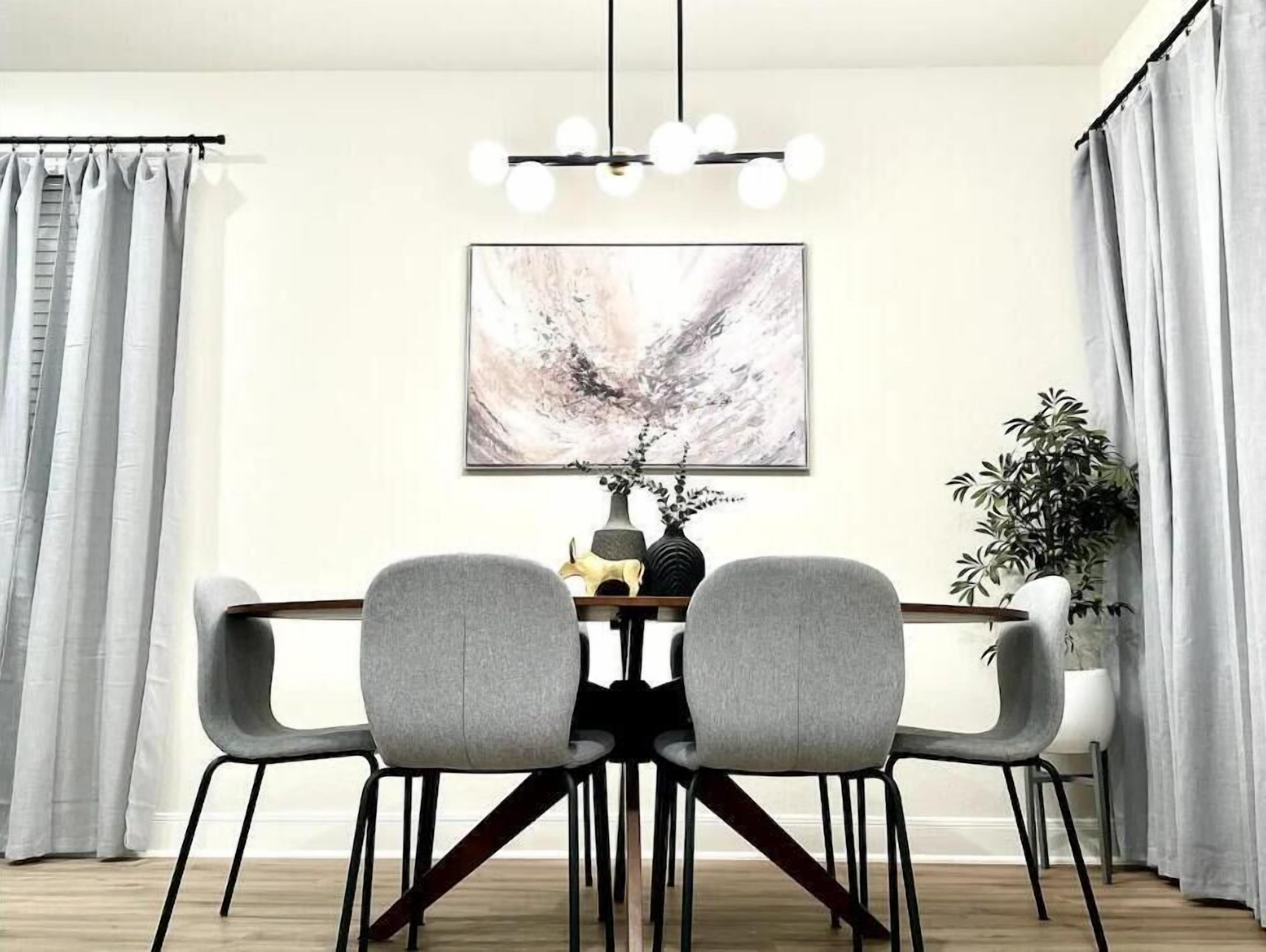 Un coin repas avec une table en bois, deux chaises grises, un lustre moderne, un tableau abstrait encadré et une plante en pot.