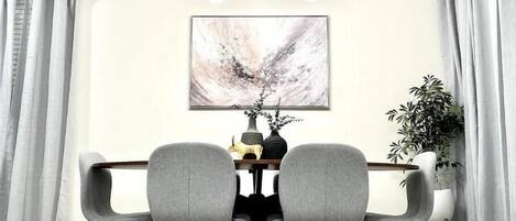 Un coin repas avec une table en bois, deux chaises grises, un lustre moderne, un tableau abstrait encadré et une plante en pot.